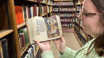 Our library assistant Alex, wearing a green jumper, holds the Book of hours open while standing in the Yorkshire room at Central Library, with rows of bookshelves in the background